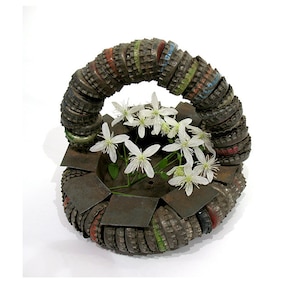 Może przedstawiać: Metalowy wazon wykonany z zakrętek od butelek, w kształcie pierścienia, z białymi kwiatami w środku.