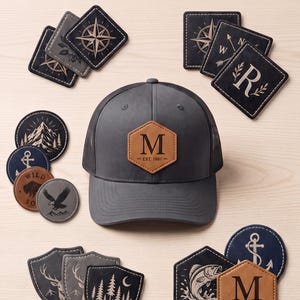 May include: A dark grey baseball cap with a tan hexagonal leather patch featuring the letter M and text EST. 1997. Surrounding the cap are various leather patches in circular, square, and shield shapes with engraved designs including compasses, animals, and trees. Text reads: YOUR LOGO ON LEATHER NO EMBROIDERY NEEDED and UV PRINTED & HEAT PRESS READY.