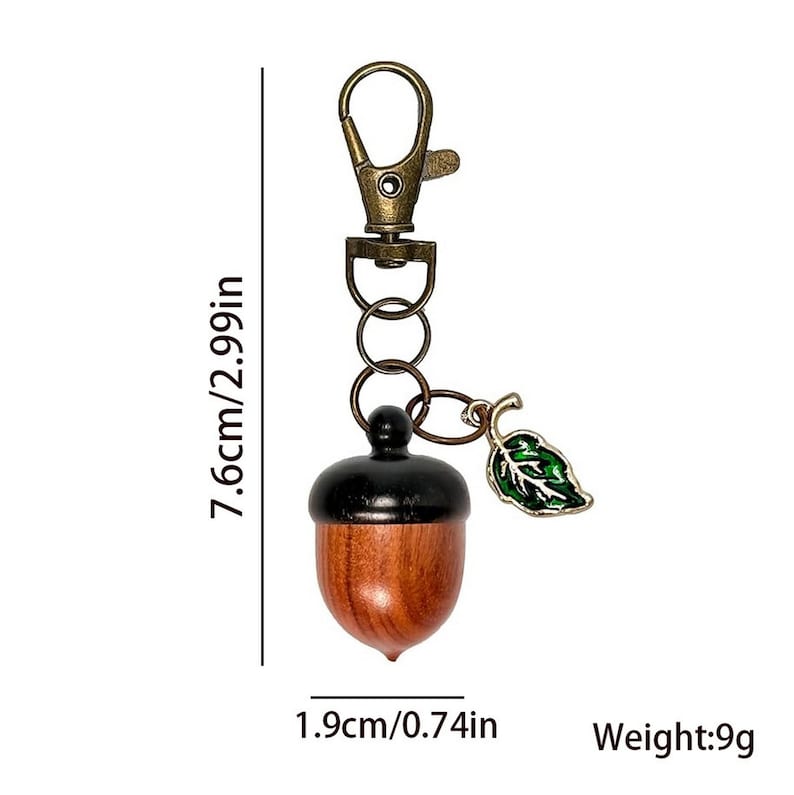 May include: A wooden acorn keychain with a bronze-colored clasp and rings. The acorn is brown with a black cap and a green and gold leaf charm. The keychain is 2.99 inches long and weighs 9g.