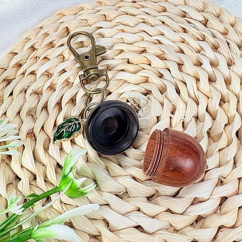 May include: A wooden acorn-shaped container with a dark brown top and a lighter brown bottom. A keychain with a gold-colored clasp and a green leaf charm is attached to the top. The items are on a woven straw mat.