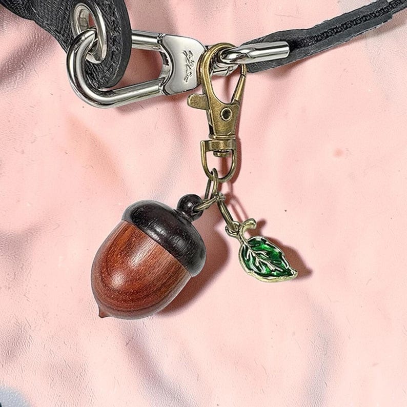 May include: A brown and black acorn charm with a green leaf charm attached to a metal clasp. The acorn is made of wood and has a dark brown cap. The leaf charm is green with gold trim. The clasp is attached to a black leather strap.
