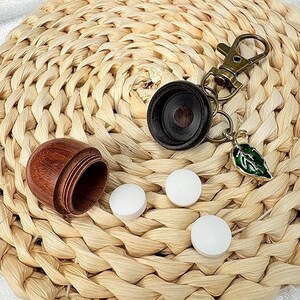May include: A wooden pill container with a screw-on lid, a black keychain with a leaf charm, and three white pills are displayed on a woven straw mat. The container is open, revealing its interior.