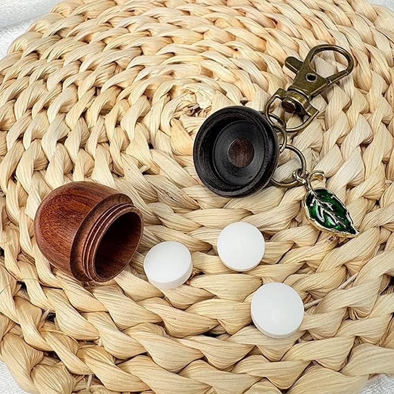 May include: A wooden pill container with a screw-on lid, a black keychain with a leaf charm, and three white pills are displayed on a woven straw mat. The container is open, revealing its interior.