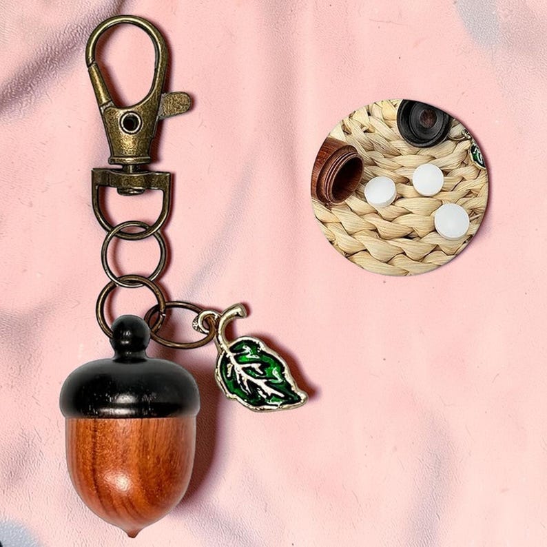 May include: A wooden acorn-shaped keychain with a black top and a bronze-colored clasp. A green and white leaf charm hangs next to the acorn. The image also shows a small wooden container and white pills on a woven surface.