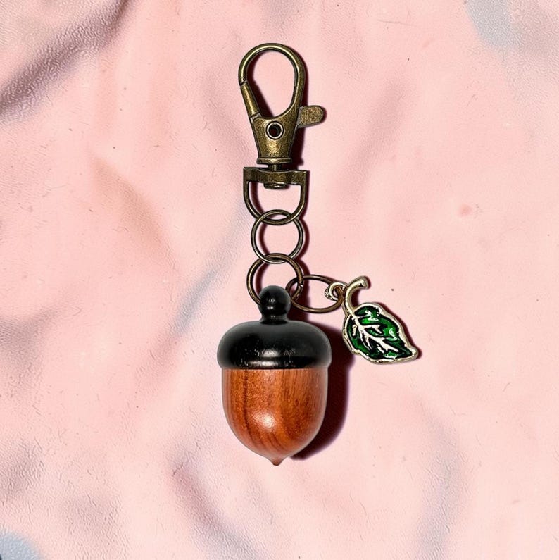May include: A wooden acorn keychain with a black cap and a bronze-colored clasp. A small green leaf charm hangs next to the acorn. The keychain is on a pink background.