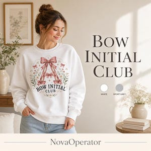 Op de afbeelding: Een wit sweatshirt met ronde hals en een afbeelding van een grote letter A gevormd door roze linten en strikken, omringd door bloemillustraties en de tekst Bow Initial Club. Het ontwerp staat centraal op de borst van het kledingstuk.
