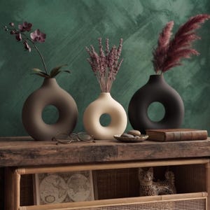 Könnte beinhalten: Drei ringförmige Keramikvasen in mattem Braun, Creme und Schwarz. Die braune Vase enthält eine dunkelviolette Orchidee, die cremefarbene Vase getrockneten Lavendel und die schwarze Vase dunkelrosa Pampasgras. Eine Brille liegt daneben.