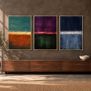 May include: A set of three vertical rectangular wall art prints in thin gold frames. The left print features teal and burnt orange tones, the center print displays deep purple and dark green, and the right print shows shades of navy blue and white.