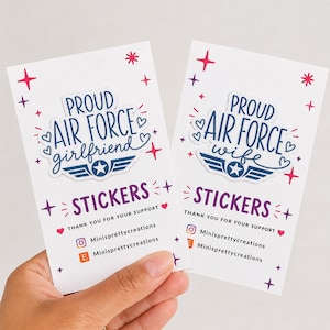Op de afbeelding: Twee witte rechthoekige displaykaarten met stickers met blauwe tekst PROUD AIR FORCE girlfriend en PROUD AIR FORCE wife. Elke sticker bevat een blauw ster-embleem met vleugels, omringd door roze en rode ster- en hartpictogrammen.
