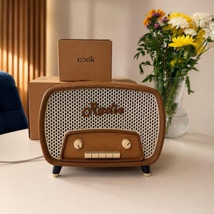 Może przedstawiać: Radio z wykończeniem imitującym brązowe drewno, białą siatkową maskownicą głośnika i napisem Radio wykonanym kursywą. Posiada dwa okrągłe pokrętła, pięć prostokątnych przycisków i cztery małe nóżki. Za nim znajduje się kartonowe pudełko z napisem nook.