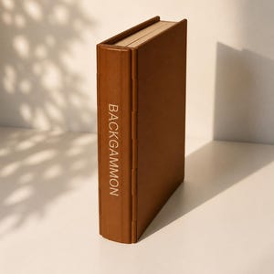 Könnte beinhalten: Ein braunes Backgammon-Spiel. Das rechteckige Spielset hat das Wort "BACKGAMMON" vertikal auf dem Buchrücken aufgedruckt. Das Set besteht aus hellbraunem Material und ist geschlossen.