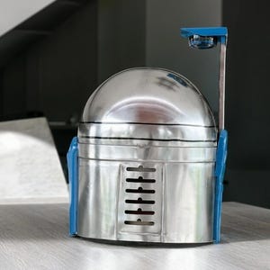 May include: A metallic silver helmet with a rounded dome top and a flat front panel featuring a vertical row of six horizontal ventilation slits. The helmet includes blue side attachments and a thin vertical arm extending upward with a blue top piece.