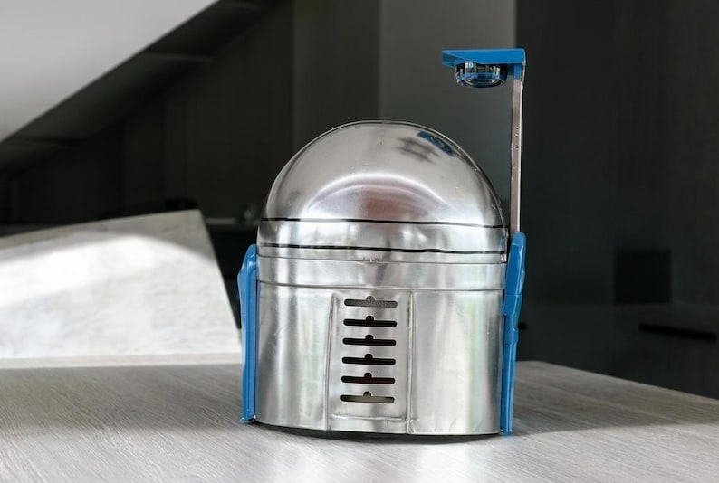 May include: A metallic silver helmet with a rounded dome top and a flat front panel featuring a vertical row of six horizontal ventilation slits. The helmet includes blue side attachments and a thin vertical arm extending upward with a blue top piece.