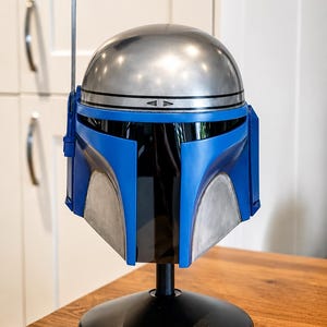 May include: A full-scale helmet featuring a metallic silver dome and a vibrant blue faceplate with a dark tinted visor. A thin metal antenna extends upward from the right side. The helmet is mounted on a black circular stand for display purposes.