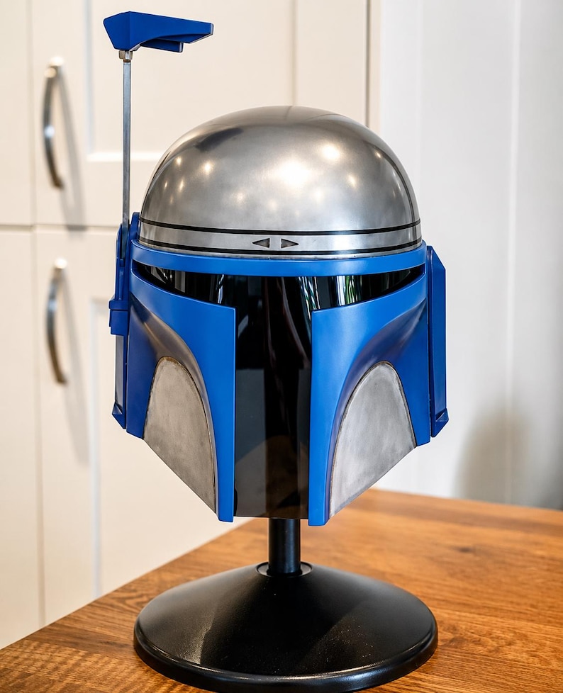May include: A full-scale helmet featuring a metallic silver dome and a vibrant blue faceplate with a dark tinted visor. A thin metal antenna extends upward from the right side. The helmet is mounted on a black circular stand for display purposes.