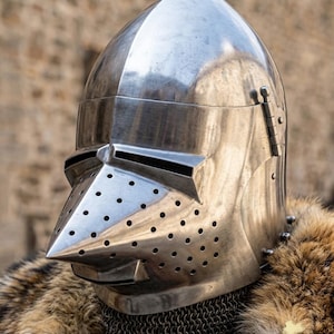 May include: A polished steel bascinet helmet featuring a pointed, perforated visor with numerous small ventilation holes and a horizontal eye slit. The helmet is worn over a metal chainmail aventail and is framed by a thick, brown faux fur collar.