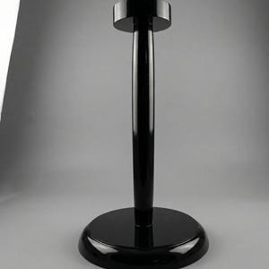 May include: A glossy black hat stand with a circular base and a cylindrical pole. The top of the stand is a smaller, circular platform. The stand is made of a shiny, reflective material.