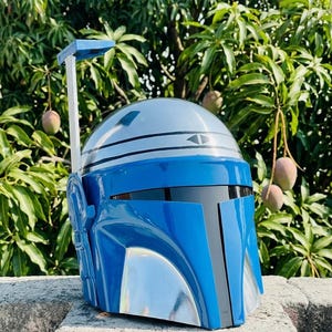 May include: A glossy blue and silver helmet featuring a smooth, rounded dome, a dark horizontal visor, and a vertical rangefinder antenna attached to the right side. The helmet has a distinct T-shaped faceplate design with metallic silver accents.