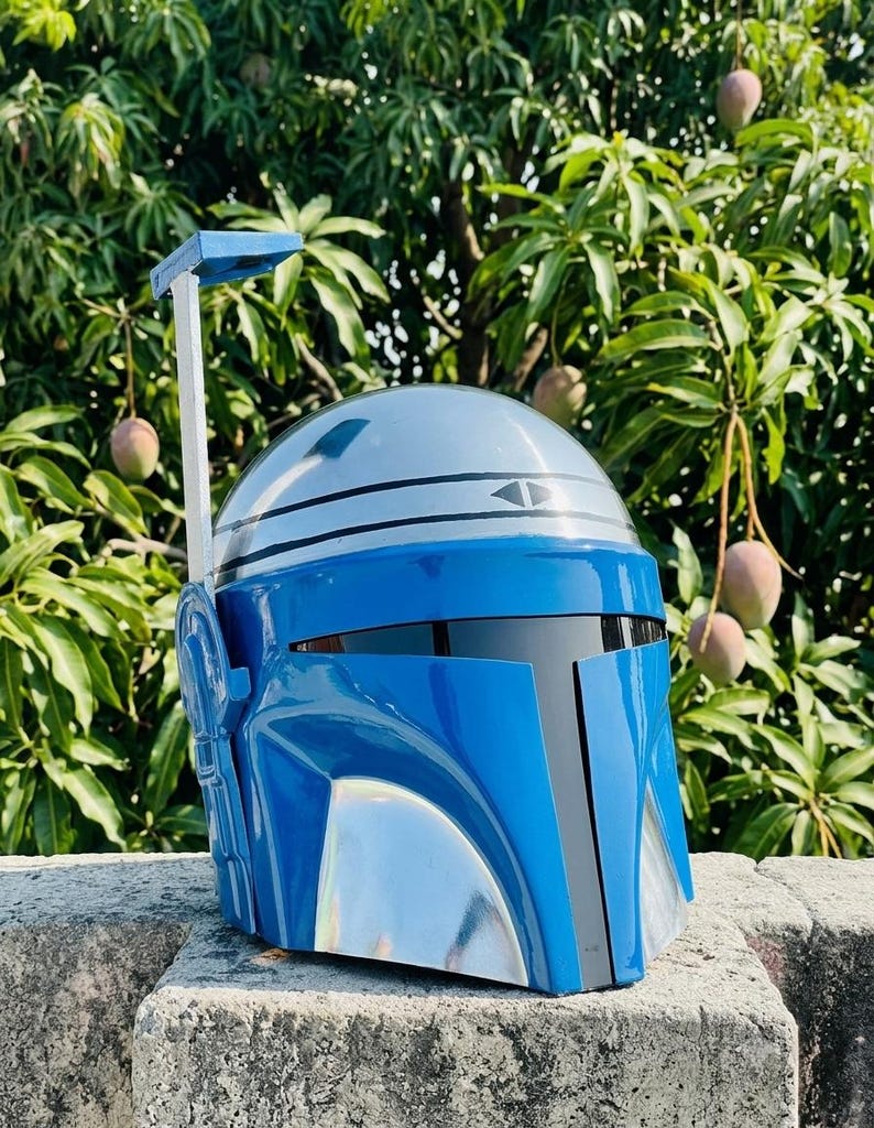 May include: A glossy blue and silver helmet featuring a smooth, rounded dome, a dark horizontal visor, and a vertical rangefinder antenna attached to the right side. The helmet has a distinct T-shaped faceplate design with metallic silver accents.