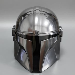 May include: A metallic silver Mandalorian helmet with a black visor. The helmet has a smooth, reflective surface with vertical ridges on top. The design features angular shapes and a sleek, futuristic aesthetic, perfect for cosplay or display.