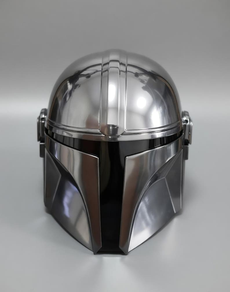 May include: A metallic silver Mandalorian helmet with a black visor. The helmet has a smooth, reflective surface with vertical ridges on top. The design features angular shapes and a sleek, futuristic aesthetic, perfect for cosplay or display.