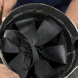 May include: The interior of a protective helmet featuring a black leather suspension liner. The liner consists of multiple overlapping leather flaps secured with thin black laces that converge at the center, providing an adjustable fit system.