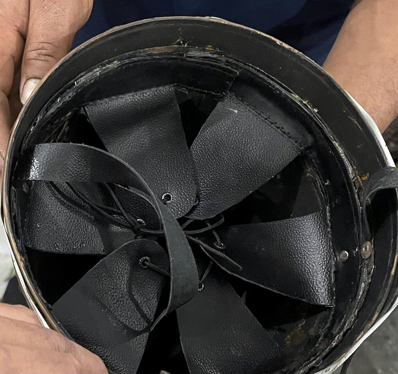 May include: The interior of a protective helmet featuring a black leather suspension liner. The liner consists of multiple overlapping leather flaps secured with thin black laces that converge at the center, providing an adjustable fit system.