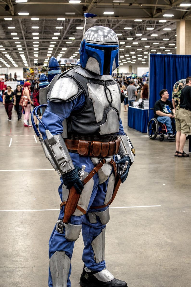 May include: A full-body Mandalorian-style costume featuring a silver and blue helmet with a T-shaped visor, silver chest and limb armor, a blue jumpsuit, a brown leather utility belt, and a jetpack with blue accents worn over the shoulders.