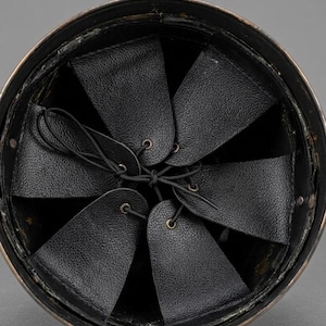 May include: An overhead view of the interior of a helmet. The helmet's interior features a series of black leather flaps, each with a small brass grommet, connected by a black drawstring. The helmet's rim is a dark metallic color.
