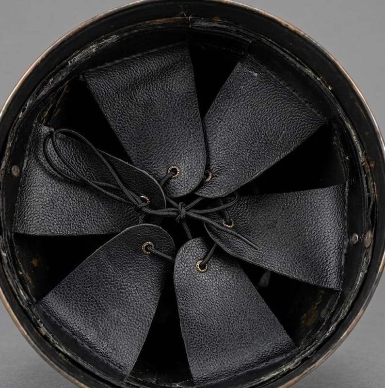 May include: An overhead view of the interior of a helmet. The helmet's interior features a series of black leather flaps, each with a small brass grommet, connected by a black drawstring. The helmet's rim is a dark metallic color.