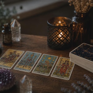 May include: A wooden table displays tarot cards with various illustrations and titles, including "The Lovers," "The Chariot," "The Star," and "The Sun." A deck of cards with a starry design, crystals, a candle, and a decorative candle holder are also present.