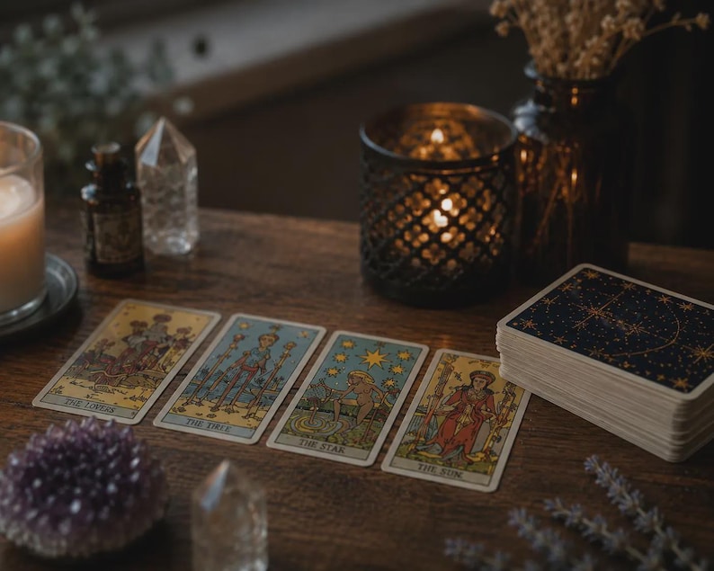 May include: A wooden table displays tarot cards with various illustrations and titles, including "The Lovers," "The Chariot," "The Star," and "The Sun." A deck of cards with a starry design, crystals, a candle, and a decorative candle holder are also present.