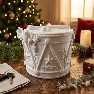 Op de afbeelding: Een matwitte decoratieve trommelhouder met een deksel waarop twee drumstokken liggen. De buitenkant is versierd met reliëfdetails, waaronder een strik, hulstbladeren, sterren en touwpatronen. Het cilindervormige object heeft een platte boven- en onderkant.