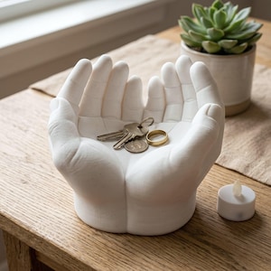 May include: A white matte ceramic trinket dish shaped like two cupped hands held together. The interior of the hands contains a silver metal key and a plain gold-colored metal ring. The sculpture has a smooth, solid finish and a rounded base.