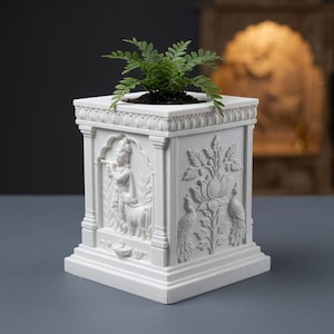 Puede incluir: Una maceta cuadrada blanca con intrincados relieves en sus lados, que incluyen una figura tocando la flauta con una vaca y un motivo floral con dos pavos reales. Una pequeña planta de helecho verde está plantada en la abertura superior.