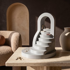 Peut inclure: Un présentoir gris clair en forme d'arche avec un design d'escalier incurvé. La structure comprend plusieurs marches, avec de petits livres miniatures placés sur les niveaux supérieurs. Un collier doré et des clés métalliques reposent sur la base.