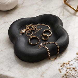 Op de afbeelding: Een zwarte, bloemvormige sieradenbak bevat gouden kettingen, ringen en een armband. De bak staat op een marmeren oppervlak, met een witte vaas en gouden geometrische decoratie op de achtergrond. De sieraden omvatten een delicate ketting met een hanger.