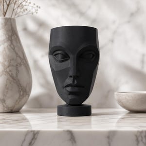 May include: A black, geometric face-shaped vase with a round base. The vase has a smooth, matte finish and is set against a marble background. A white marble vase with dried flowers is to the left.