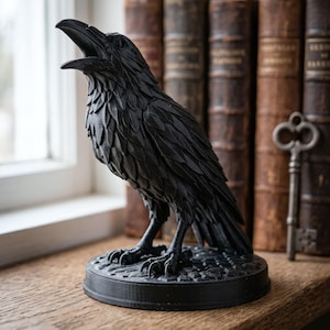 May include: A matte black raven figurine standing on a circular base with a textured surface. The bird is depicted with its beak open, showing detailed feather textures across its body, wings, and head, captured in a static, upright pose.