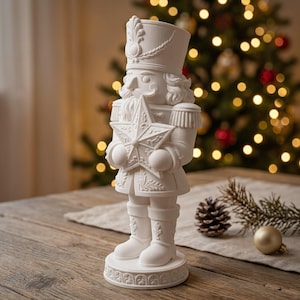 Pode incluir: Uma estatueta de quebra-nozes em branco mate sobre uma base circular. O quebra-nozes usa um chapéu alto com um detalhe de pena, um uniforme com dragonas e segura uma grande estrela de cinco pontas com padrões em relevo nas mãos.