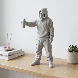 Puede incluir: Una figura monocromática de color gris claro que representa a una persona con sudadera con capucha y cubrebocas, pantalones cargo y zapatillas. La figura está de pie, extendiendo un brazo para sostener un pequeño bote de spray cilíndrico.