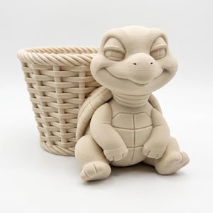 Puede incluir: Una maceta de cerámica beige claro con forma de tortuga y una cesta tejida. La tortuga tiene una cara sonriente y está sentada con las piernas extendidas. La cesta es rectangular y tiene un diseño tejido.