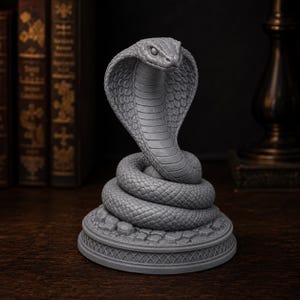 May include: A grey, detailed figurine of a cobra snake with its hood flared, coiled atop a circular base featuring a textured, stone-like surface and a decorative lattice pattern around the lower edge, captured in a neutral, monochromatic finish.