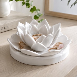 以下が含まれることがあります： 円形に重なった花びらを持つ、白い陶器製の蓮の花の形をしたジュエリーディッシュ。中央と外側の花びらには、様々なゴールドトーンの指輪や繊細なジュエリーが置かれており、滑らかな円形の土台の上に配置されています。