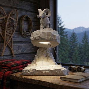 May include: A white table lamp featuring a sculpted ram standing on a rocky mountain base. The lamp has a circular light source integrated into the underside of the upper rock formation, casting a downward glow onto the lower rock structure.