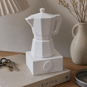May include: A white, faceted moka pot sculpture resting on a square base featuring a single coffee bean relief. The geometric coffee maker has a lid, a handle, and a spout, all rendered in a matte, solid white finish with clean, sharp edges.