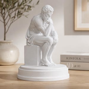 Puede incluir: Una escultura de color blanco mate de una figura sentada y barbuda en pose contemplativa, con la barbilla apoyada en una mano. La figura se asienta sobre un bloque cuadrado encima de una base circular escalonada, con un acabado blanco liso.