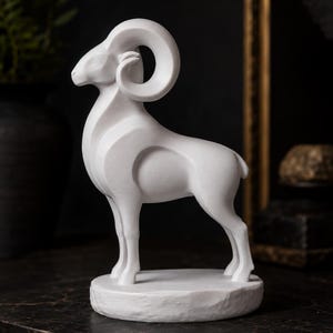 Könnte beinhalten: Eine mattweiße Skulptur eines Widders, der auf einem runden Sockel steht. Die Figur zeichnet sich durch glatte, stilisierte Kurven, ein markantes, eingerolltes Horn und einen definierten Körperbau aus. Der Widder ist im Profil nach links ausgerichtet.