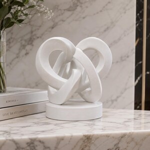 May include: A matte white sculptural object featuring interconnected, curved, loop-like forms mounted on a circular base. The sculpture has a smooth, solid finish and is positioned next to two stacked books with the text KINFOLK and LIVE BEAUTIFULLY.