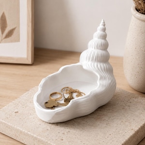 Puede incluir: Un cuenco de cerámica blanca con forma de concha marina y parte superior en espiral. El interior de la concha sirve como recipiente poco profundo, que actualmente contiene un anillo de metal dorado y una pequeña llave, mostrando su uso de almacenaje.